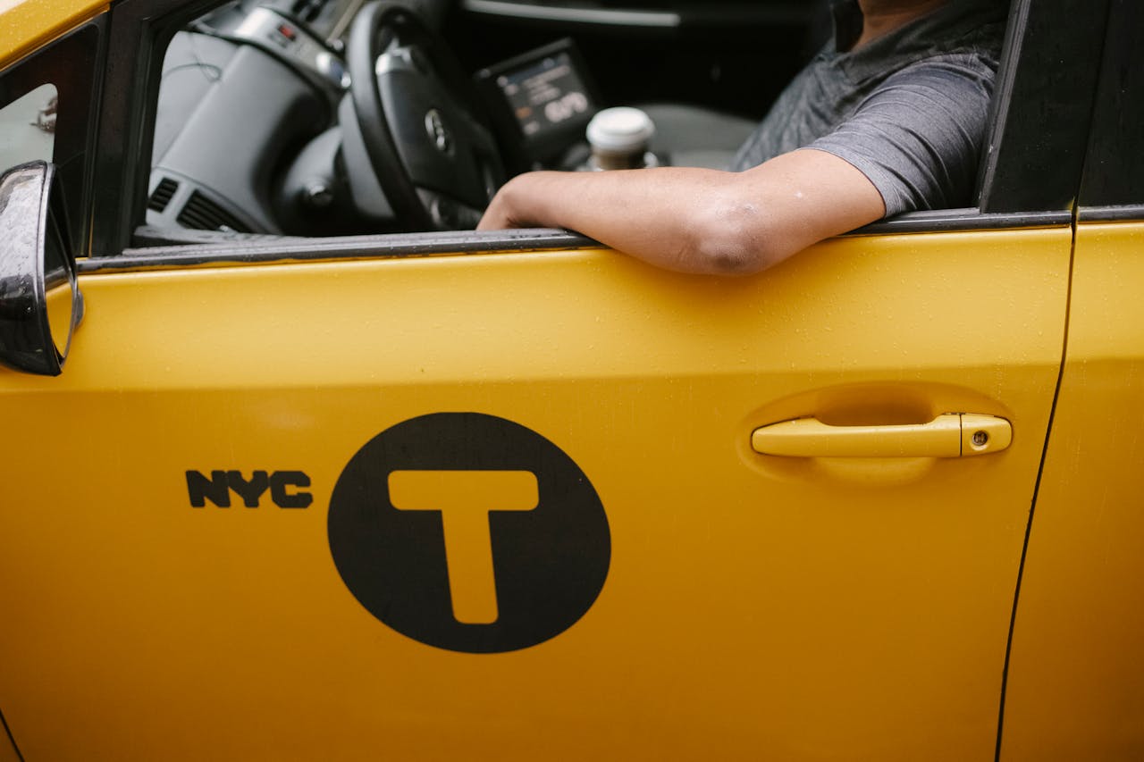 hero-img-02 A close-up view of a yellow NYC taxi with a driver resting an arm on the window, showcasing urban transportation.